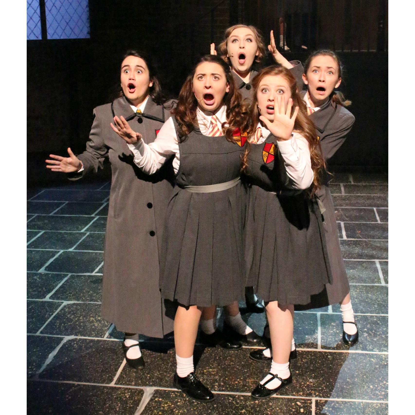 Five performers in gray school uniforms pose with surprised open mouths and raised hands on stage. Gemma Evans - Daisy Pulls it off 