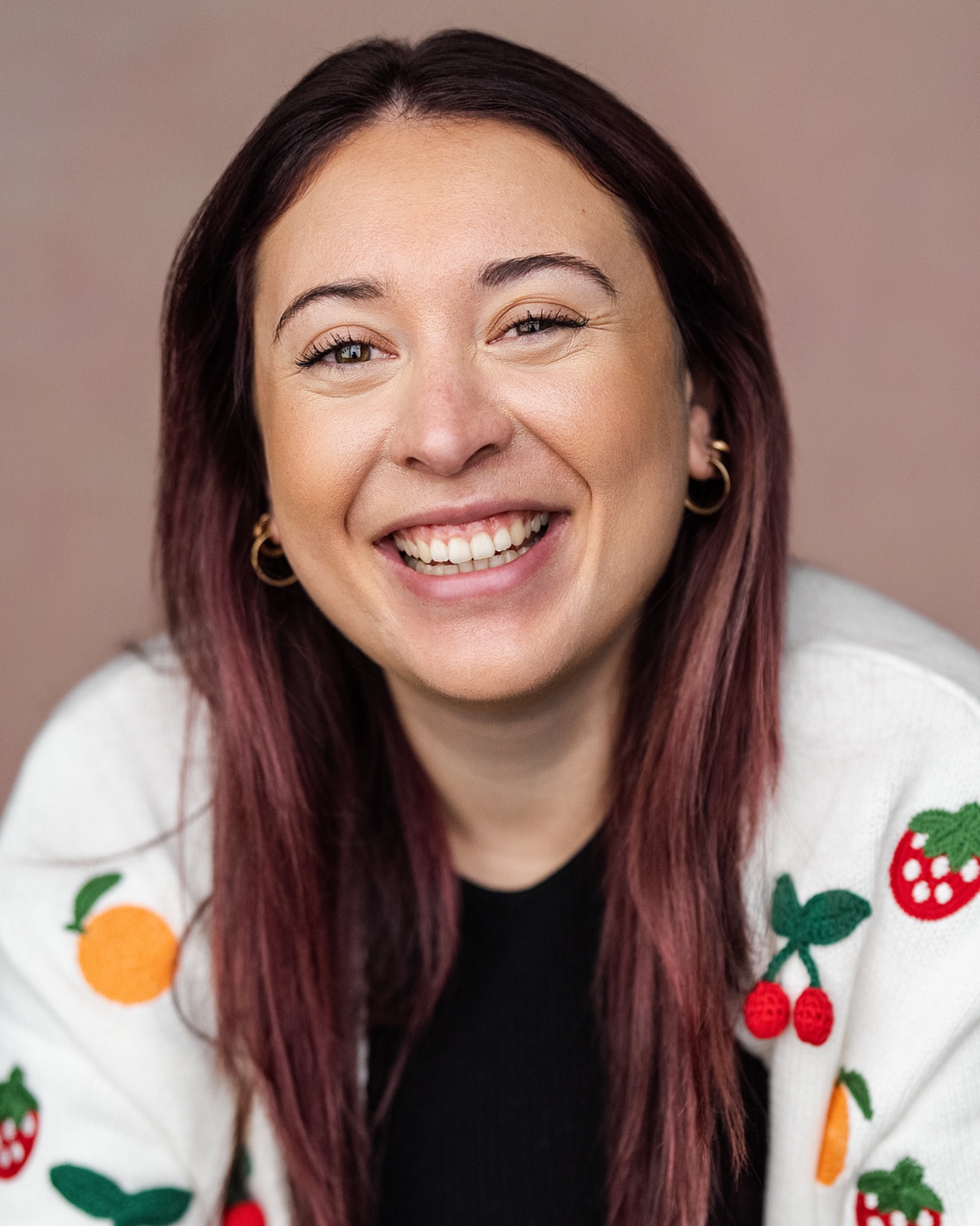 Smiling woman with burgundy hair, hoop earrings, and a festive sweater, close-up portrait against neutral background. Music and drama lessons in Newham